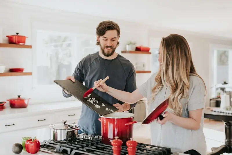 A couple cooking together in a bright white kitchen, adding chopped ingredients to a red pot on a gas stove.